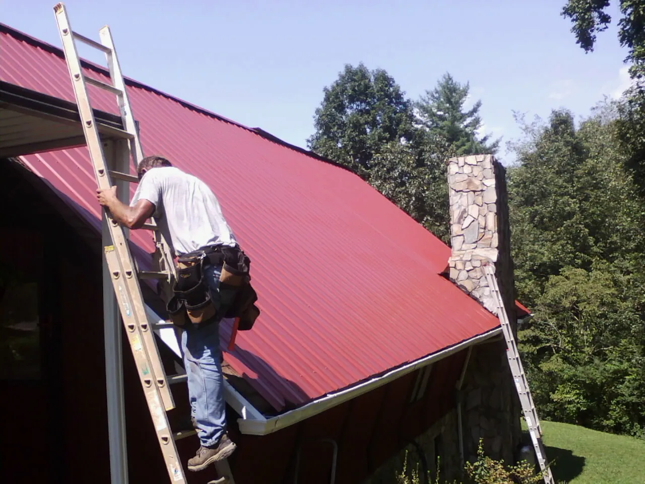 Roofer on a ladder beside a red metal roof and stone chimney — Soffit & Fascia Repair by Roofing Brothers in Willard